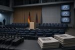 Leerer Plenarbereich mit unbesetzten Stühlen, Rednerpult und ausgeschalteten Bildschirmen. Im Vordergrund Mikrofonständer und ungelesene Zeitungen.