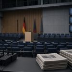 Leerer Plenarbereich mit unbesetzten Stühlen, Rednerpult und ausgeschalteten Bildschirmen. Im Vordergrund Mikrofonständer und ungelesene Zeitungen.